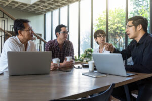 紙コップのコーヒーを飲みながら、パソコンを開き話をする男女四人