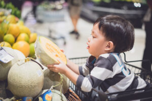 スーパーマーケットでメロンを手に取る買い物カートに乗った男の子
