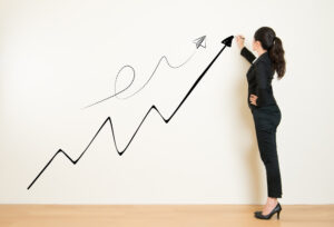 パンツスーツで束ねた髪の女性が白い壁に向かって、上昇する矢印を描く様子