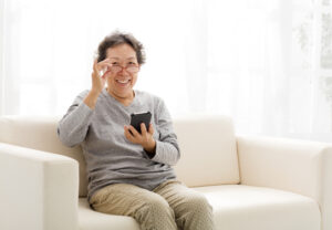 眼鏡を左手で持ち上げながらスマホを手にしてソファに座る笑顔のおばあさん